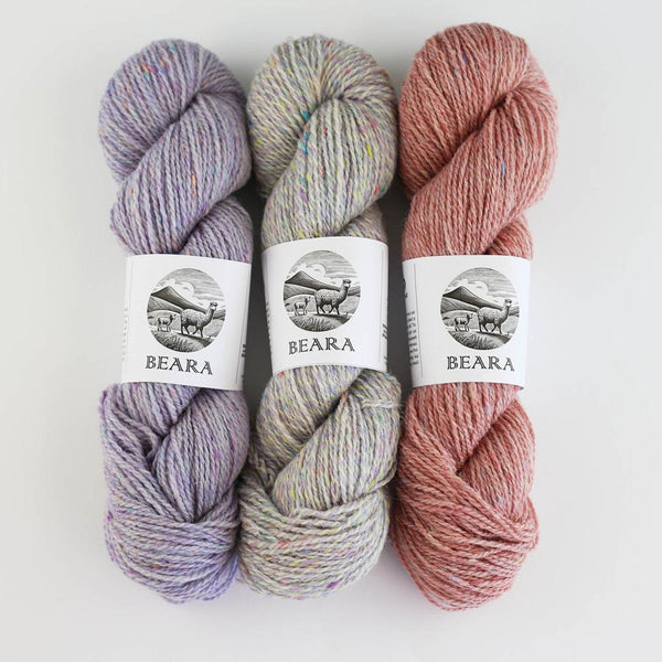 Three skeins of Beara Irish wool and UK alpaca tweed dk, worsted or aran weight yarn by Hey Mama Wolf in purple, grey, and pink on a white background. Donegal Tweed.