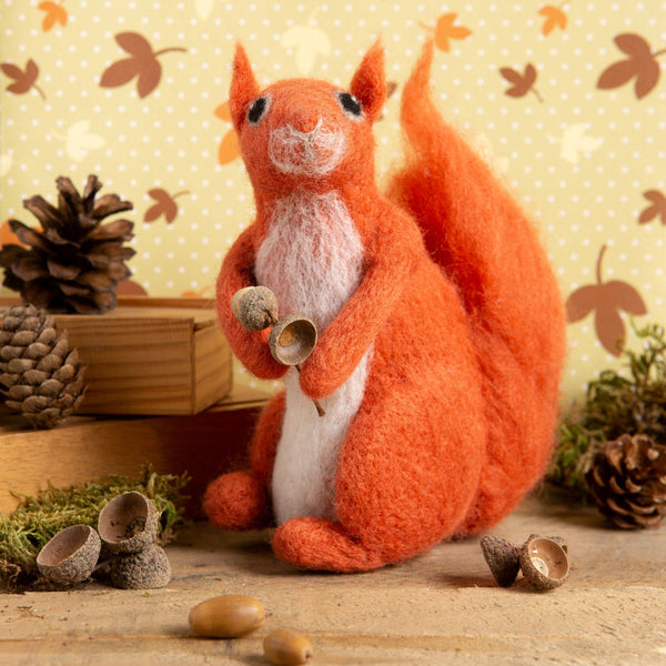 needle felted red squirrel on a table styled with acorns and pinecones in front of a background with a spots and autumn leaves design