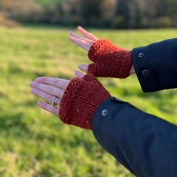 Burnt orange knitted burnt orange wrist warmers in chunky yarn on hands outstretched in front of a blurred view of fields and hills in autumn.